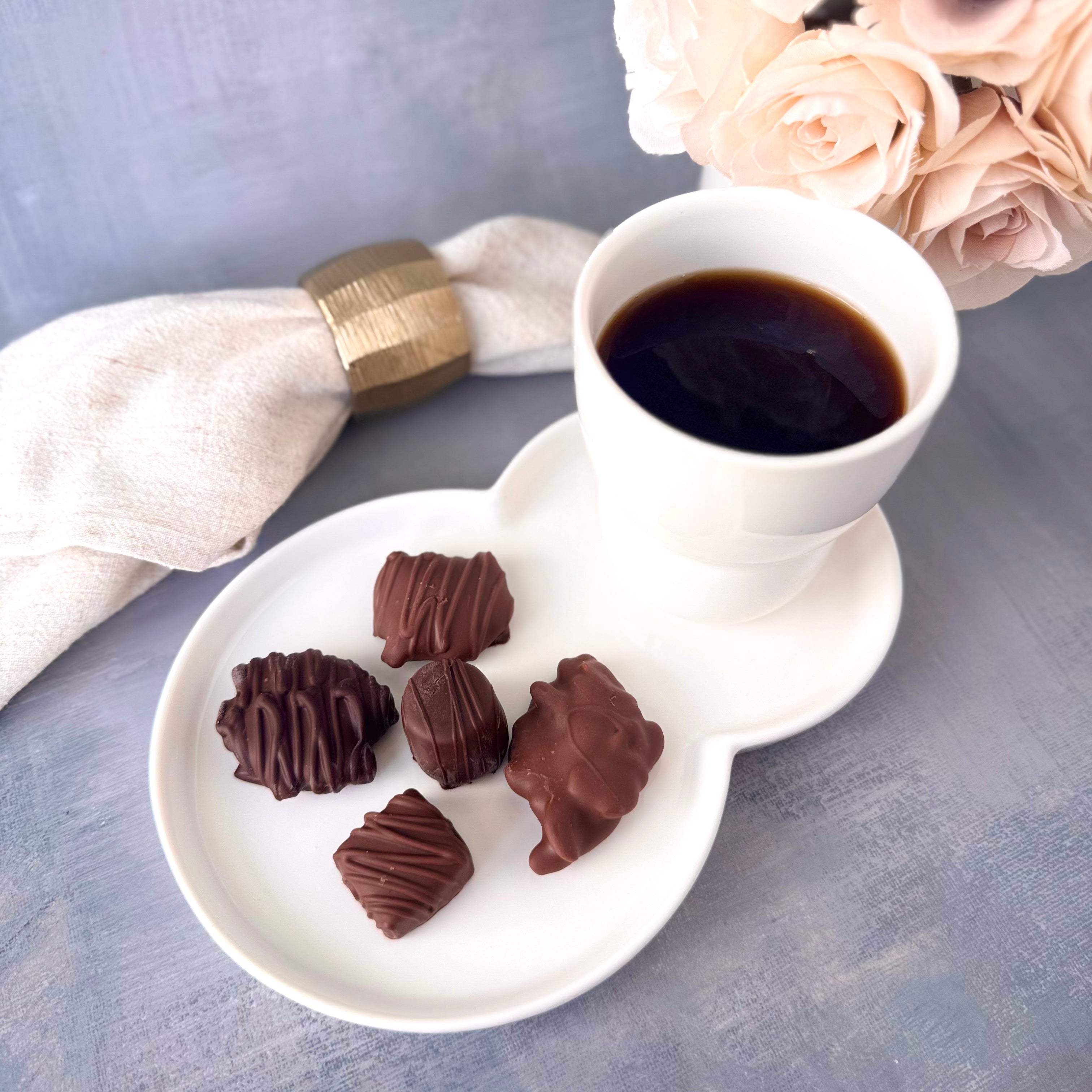 White cup of coffee on a tray with chocolate truffles and a bouquet of flowers on a gray surface.